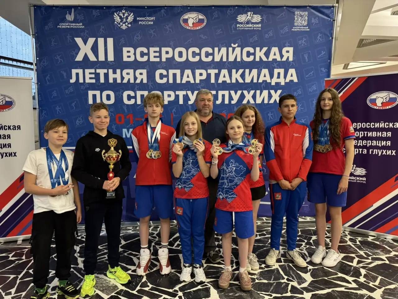 Группа спортсменов с медалями и кубком на фоне баннера всероссийской спартакиады по спорту глухих